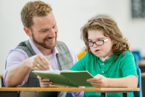 A child with a tracheostomy in a school setting -reading a book, accompanied by a smiling adult.